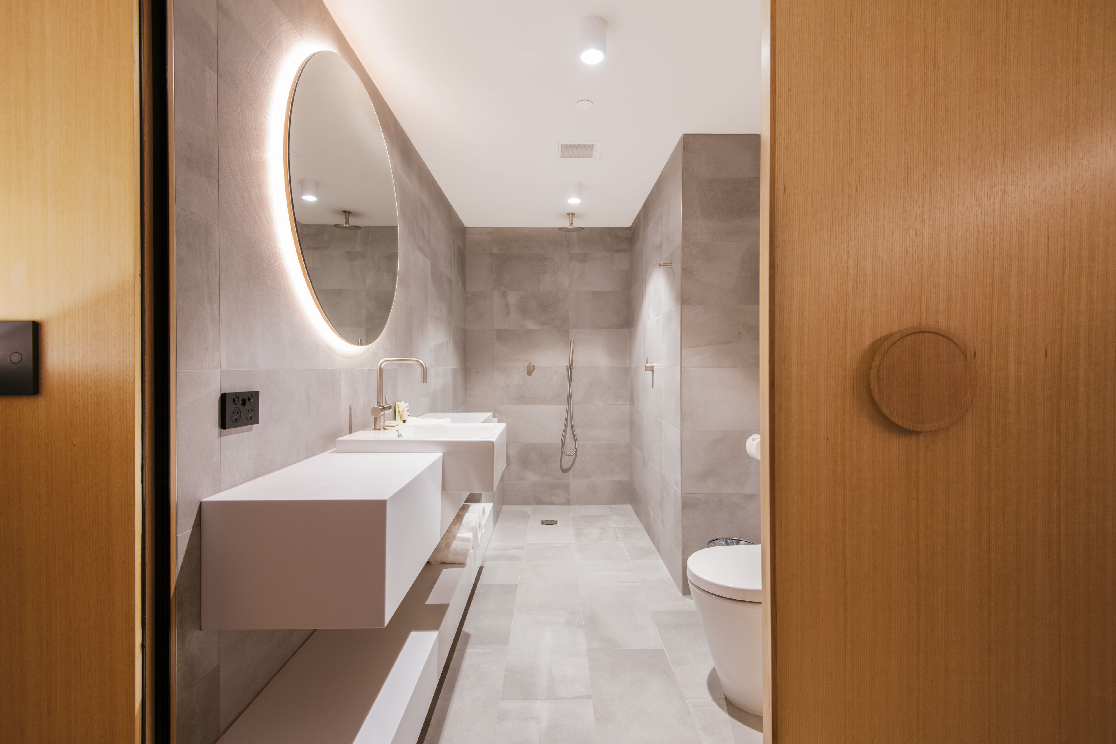 Un baño moderno con puerta de madera, un espejo grande, dos lavamanos, un inodoro y una zona de ducha.