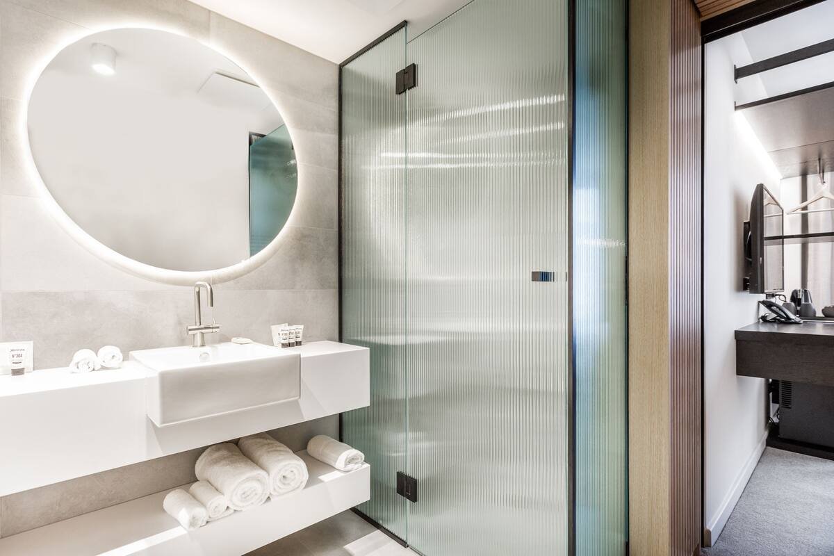 Une salle de bain moderne avec un miroir rond, un lavabo blanc et une cabine de douche en verre.