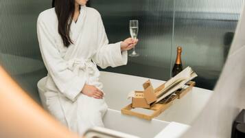 A person in a white bathrobe holding a champagne glass, seated at a table with a bottle and a wooden tray.