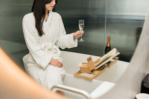 A person in a white bathrobe holding a champagne glass, seated at a table with a bottle and a wooden tray.