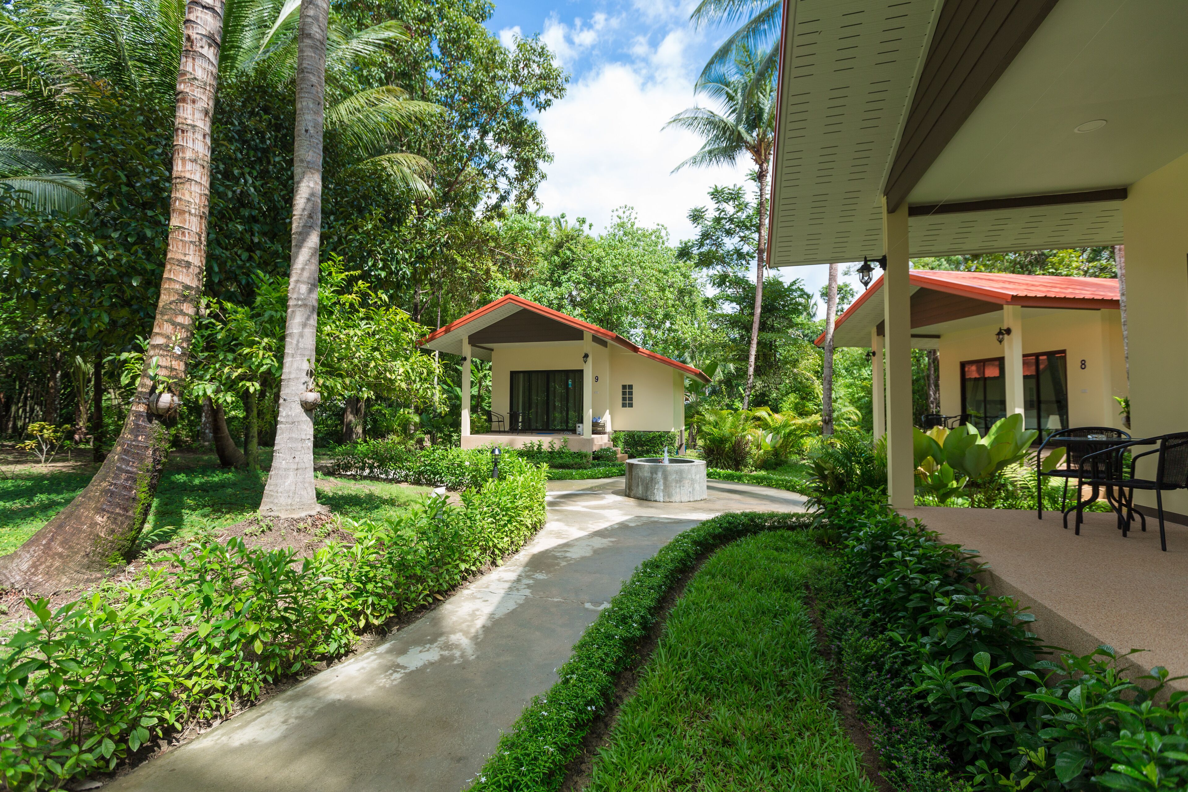 A tropical resort with a paved pathway, palm trees, and two buildings with red roofs.