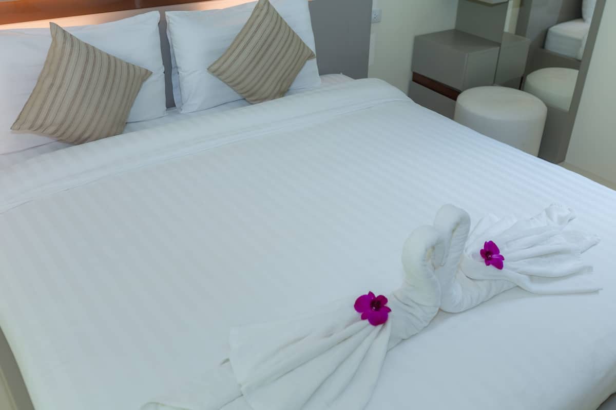 Een net opgemaakt bed met witte lakens, twee kussens en twee handdoeken gevouwen in de vorm van een zwaan, versierd met paarse bloemen.