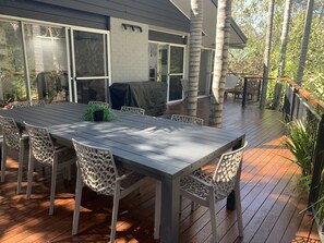 An outdoor deck with a dining table and chairs, surrounded by trees and a house. - Family Friendly Beach House - 2min walk to Beach, Bushwalks & Open Fireplace (Vincentia)
