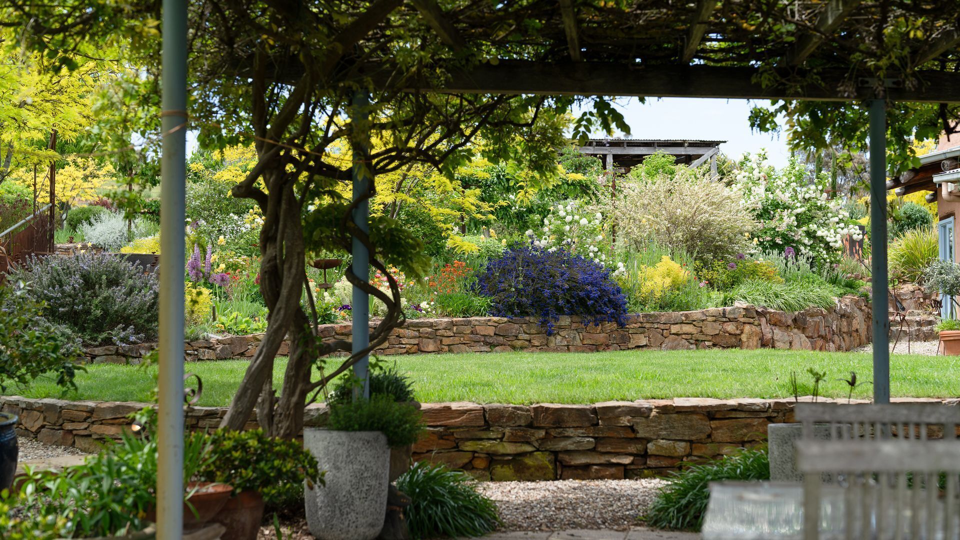 Un jardin avec un mur en pierre, diverses plantes et une structure en forme de pergola.