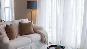 Un sofá blanco con cojines marrones, una mesita redonda con una bandeja y cortinas de encaje.