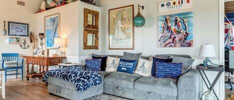 A cozy living room with a grey sofa, a wooden coffee table, and a blue patterned rug. There are wall decorations including a map, a framed painting, and a welcome sign.