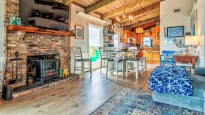 A cozy living room with a brick fireplace, wooden beams, and a dining area with a kitchen in the background. - Sea Dog- Pool, Crows Nest, Great View! 3 Bedrm,/3 Bath, Pet Friendly, Sleep 11 (Dauphin Island)