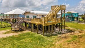 A wooden deck structure with stairs leading to a platform, surrounded by grassy areas and buildings with white and blue exteriors. - Sea Dog- Pool, Crows Nest, Great View! 3 Bedrm,/3 Bath, Pet Friendly, Sleep 11 (Dauphin Island)