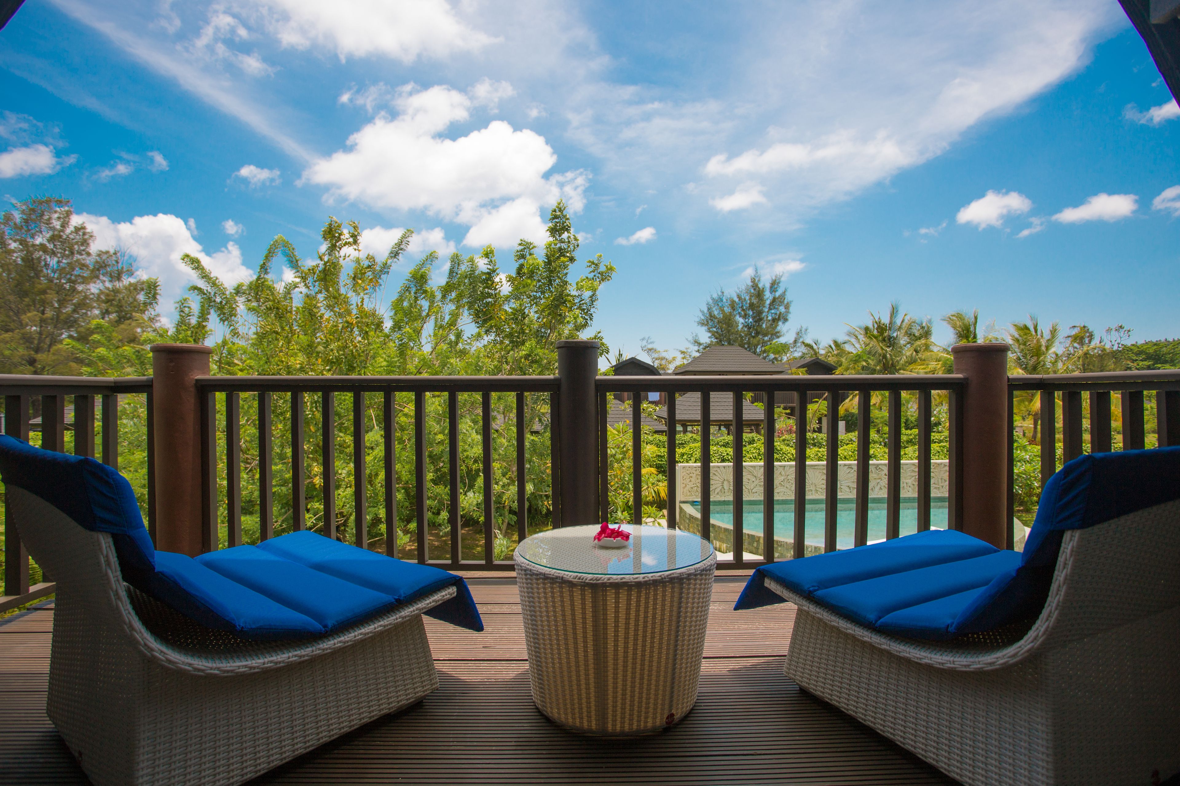 Terraza con sillones de mimbre y una mesa con cubierta de vidrio, con vista a una piscina y exuberante vegetación.