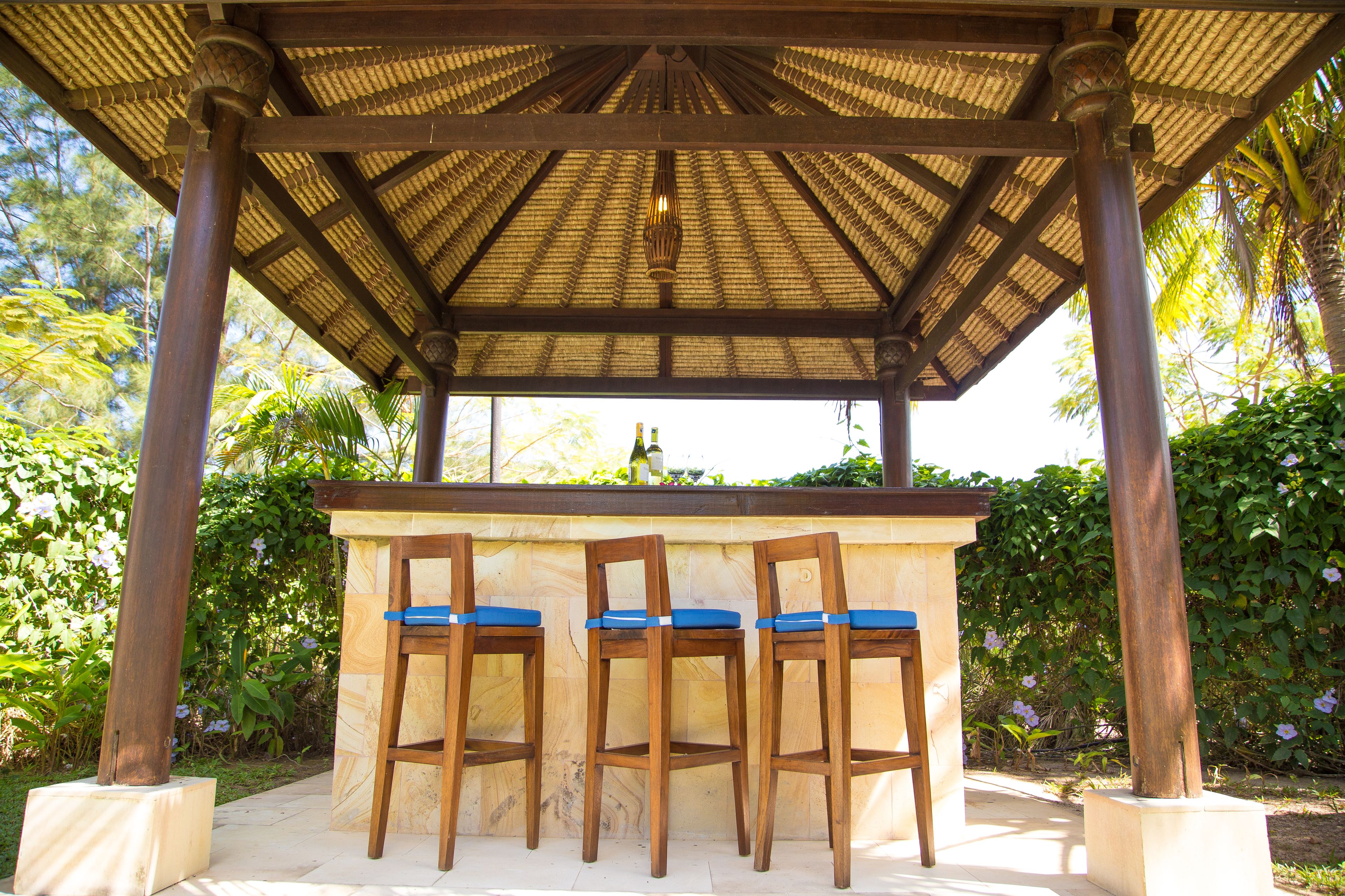 A wooden bar with three chairs under a thatched roof structure, surrounded by greenery.