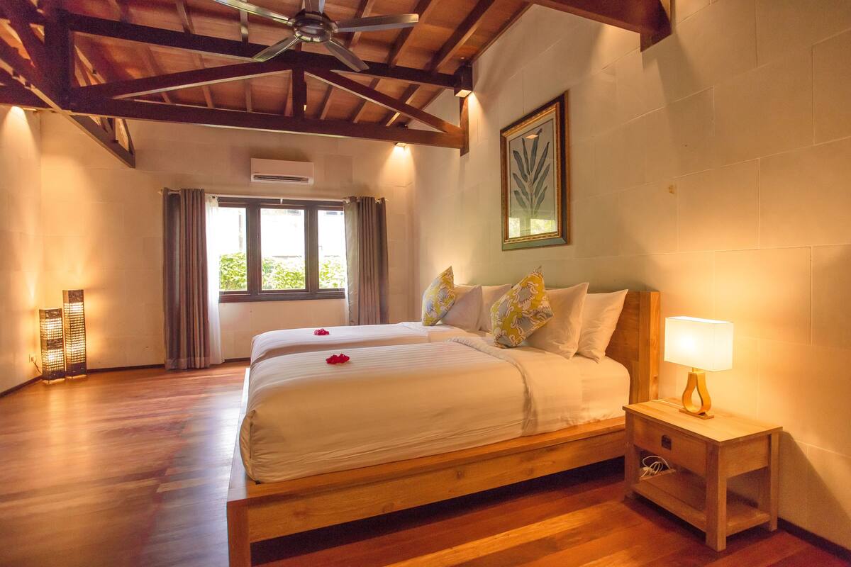 A bedroom with a wooden bed, a nightstand with a lamp, a ceiling fan, and a window with curtains.