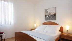 Ein ordentlich bezogenes Bett mit Holz-Kopfteil, zwei Nachttische mit Lampen, ein Fenster mit durchsichtigen Vorhängen und ein Bild an der Wand.