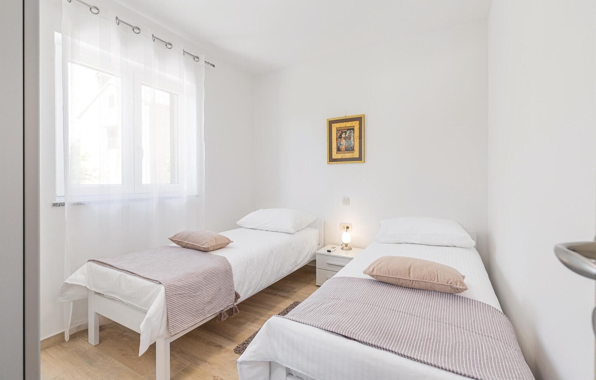 A bedroom with two single beds, a bedside table with a lamp, a window with sheer curtains, and a framed picture on the wall.