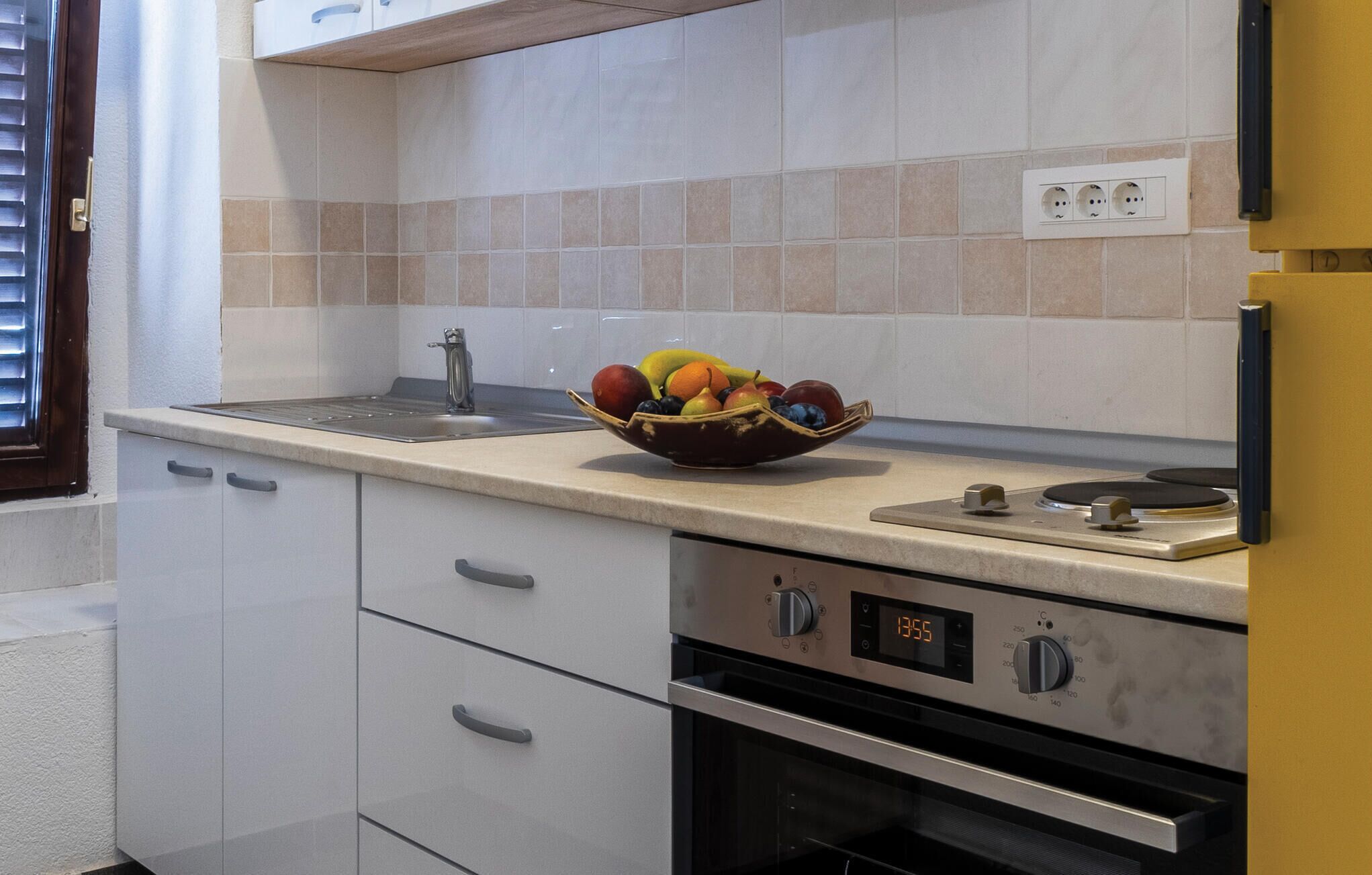 A kitchen with a stainless steel oven, a digital clock displaying 13:55, and a fruit bowl on the counter.