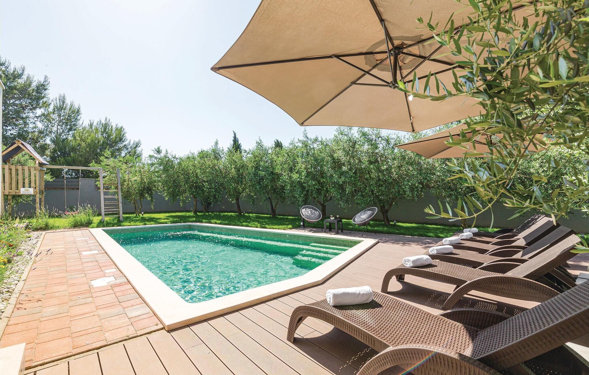 Una piscina rectangular con una cubierta de madera que la rodea, dos sillones de mimbre y un gran paraguas que da sombra.
