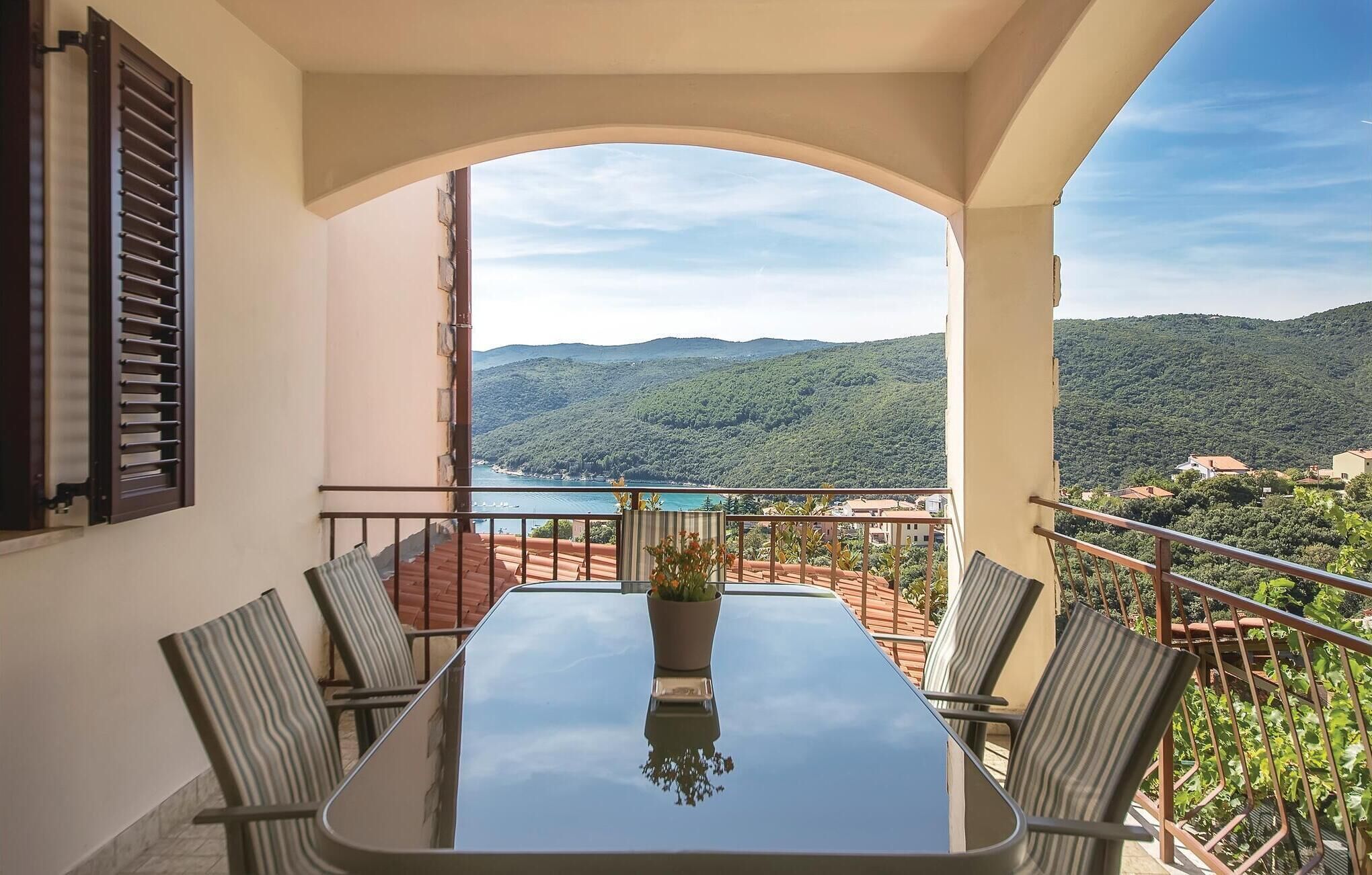 A balcony with a table and chairs, overlooking a mountainous landscape and a body of water.