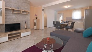 A modern living room with a TV, sofa, and glass coffee table. There is a dining area with chairs and a table. The kitchen is visible with a fridge and cabinets. - Stunning home in Vodnjan (Vodnjan)