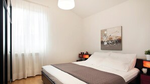 Cama bem arrumada com roupa de cama branca e castanha, mesa de cabeceira com abajur e pintura abstrata na parede.