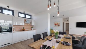 A modern kitchen with white cabinets, a built-in oven, and a wooden countertop. There is a dining area with a wooden table set for two, a sofa, and a TV mounted on the wall. - Luxurious home in Pula (Pula)