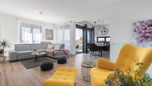 A modern living room with a grey sofa, yellow ottoman, and a dining area with a table and chairs. - Luxurious home in Pula (Pula)