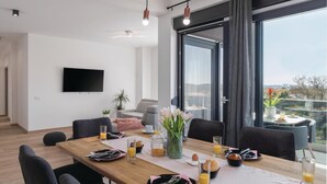 A modern dining area with a wooden table set for breakfast, large windows with a view, and a flat-screen TV mounted on the wall. - Luxurious home in Pula (Pula)