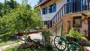 Une maison traditionnelle avec un balcon, une charrette en bois et un jardin agrémenté d’un arbre et de fleurs.