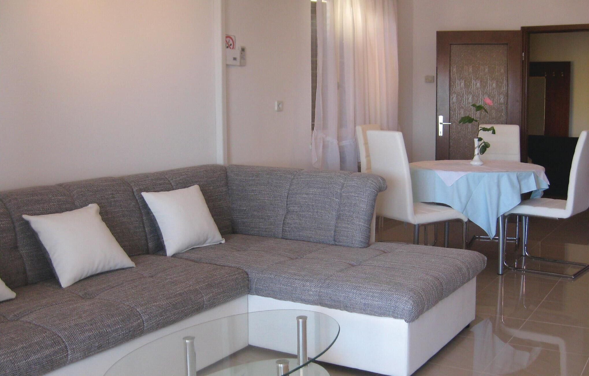 A living room with a grey sectional sofa, a glass coffee table, a dining area with a table and chairs, and a door with a plant.
