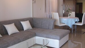 A living room with a grey sectional sofa, a glass coffee table, a dining area with a table and chairs, and a door with a plant. - Nice apartment in Rabac with house sea view (Rabac)
