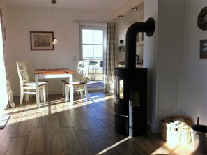 A cozy living room with a wooden table and chairs, a black wood stove, and a large window with a view. - Apartment EC - House beach runner (Dranske)
