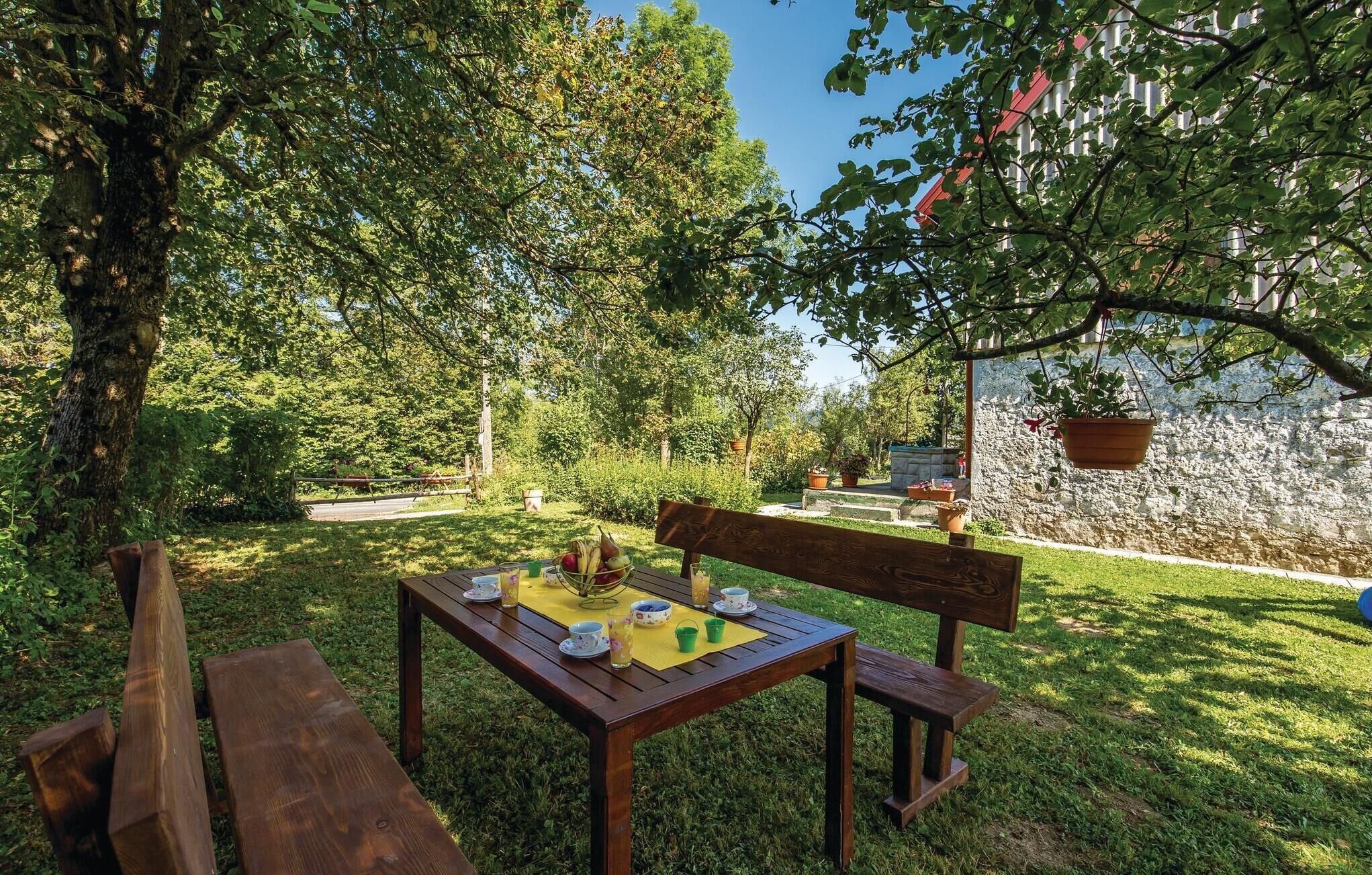 A wooden table with a yellow placemat, surrounded by benches, set in a lush garden with trees and a stone wall in the background.