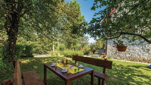 Een houten tafel met een gele placemat, omringd door banken, gelegen in een weelderige tuin met bomen en een stenen muur op de achtergrond.