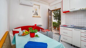 Une cuisine compacte avec des meubles blancs et un plan de travail rouge, un coin repas avec un canapé rouge et une table dressée pour deux personnes, et une plante en pot sur la table.