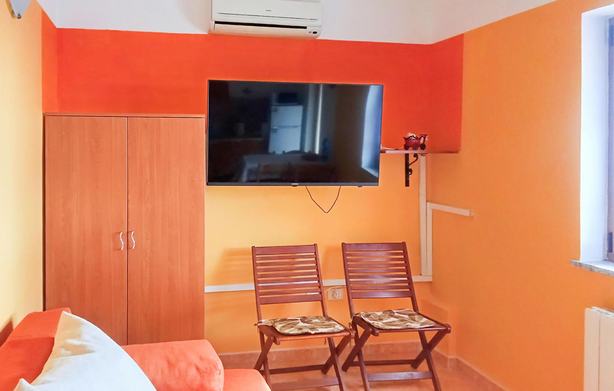 A room with orange walls, a TV mounted on the wall, a wooden wardrobe, and two wooden chairs.