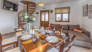 Un coin repas avec une table en bois dressée pour quatre personnes, entourée de chaises. On aperçoit un escalier au fond, menant à l’étage supérieur.