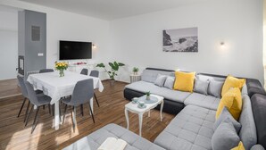 Ein modernes Wohnzimmer mit grauem Sofa, einem Esstisch mit Stühlen, einem Flachbildfernseher und einem Bild an der Wand.