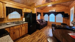 A cabin interior with wooden walls and ceiling, a kitchen with granite countertops, a black refrigerator, a brown sofa, a wooden dining table, and a TV mounted on the wall.