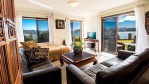 A spacious living room with a large sofa, a coffee table, and a television. There is a sliding glass door leading to a balcony with a view of the city and water. - Waterfront on Manly Harbour (Manly)