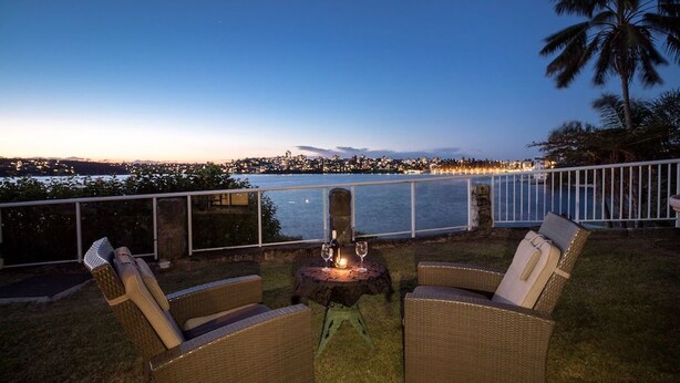 An outdoor seating area with wicker chairs and a table, overlooking a body of water with city lights in the background. - Waterfront on Manly Harbour (Manly)