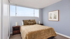 A bedroom with a large window, a bed with a beige quilted bedspread, two bedside tables with lamps, and a framed picture on the wall. - 713 at the Beach (Manly)