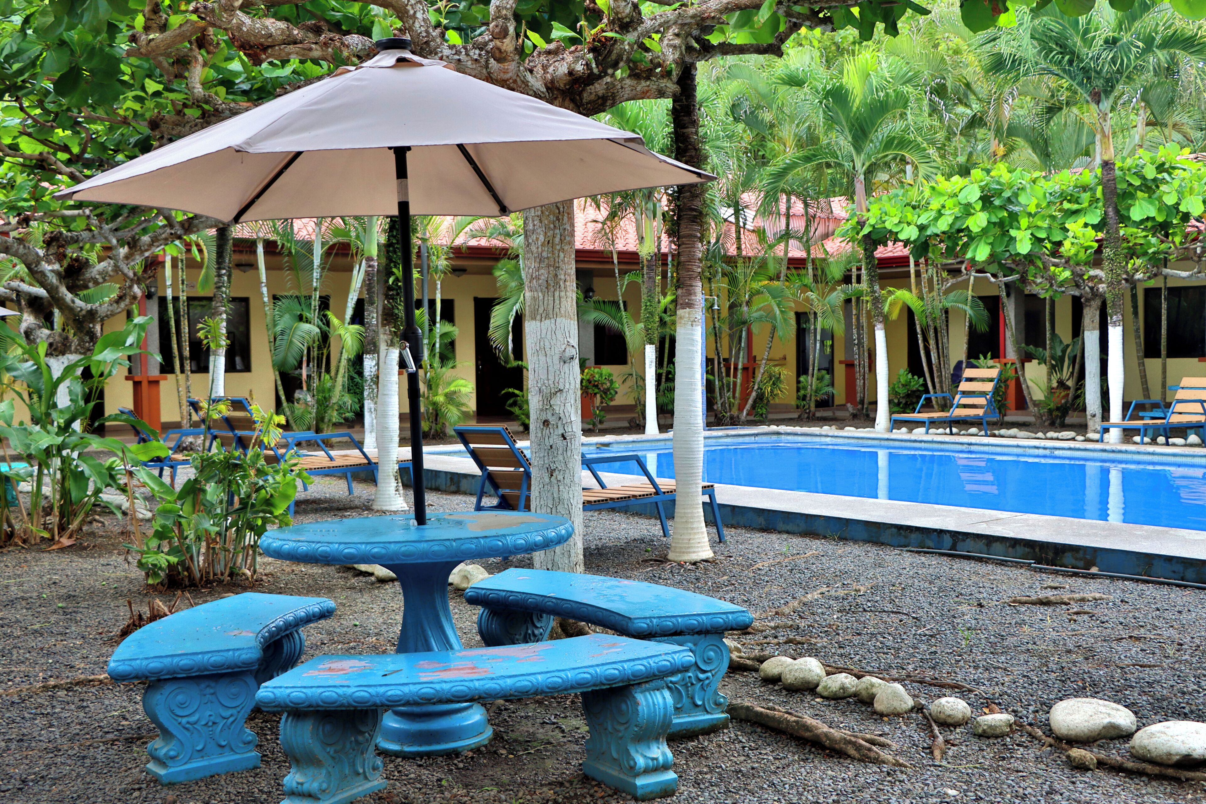 A poolside area with a table and chairs, surrounded by palm trees and a building with a red roof.