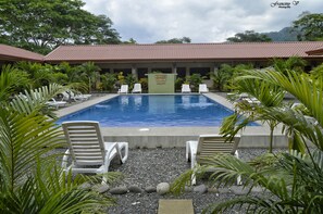 Una piscina con sillas de descanso, rodeada de plantas tropicales y un edificio con techo de tejas.