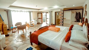 A spacious bedroom with a large bed, a wooden wardrobe, a seating area with a chair and table, and a view through a glass door to an outdoor area. - Ingagi Park View Lodge (Kinigi)