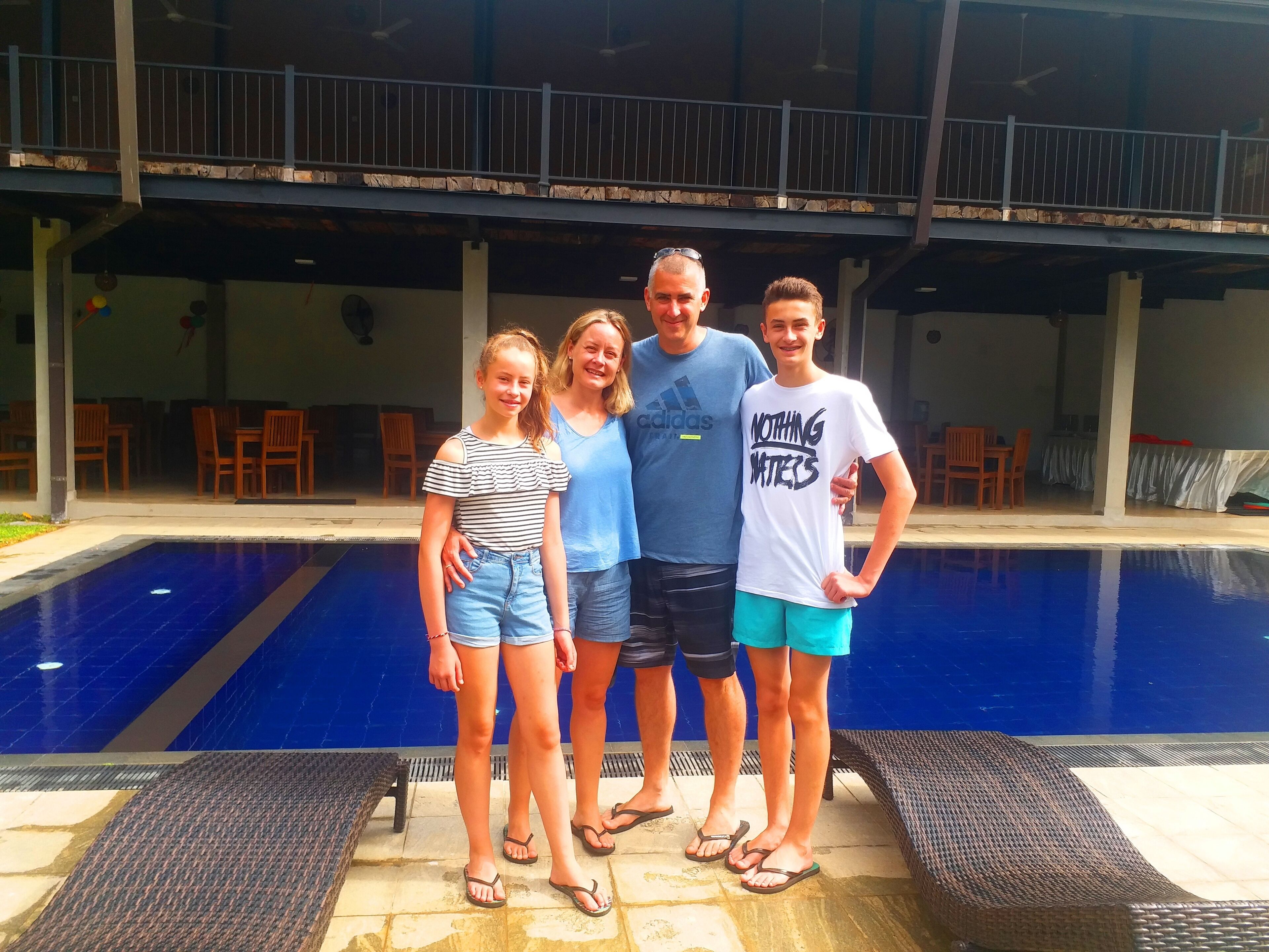 A family of four standing by a swimming pool with a covered patio area in the background.