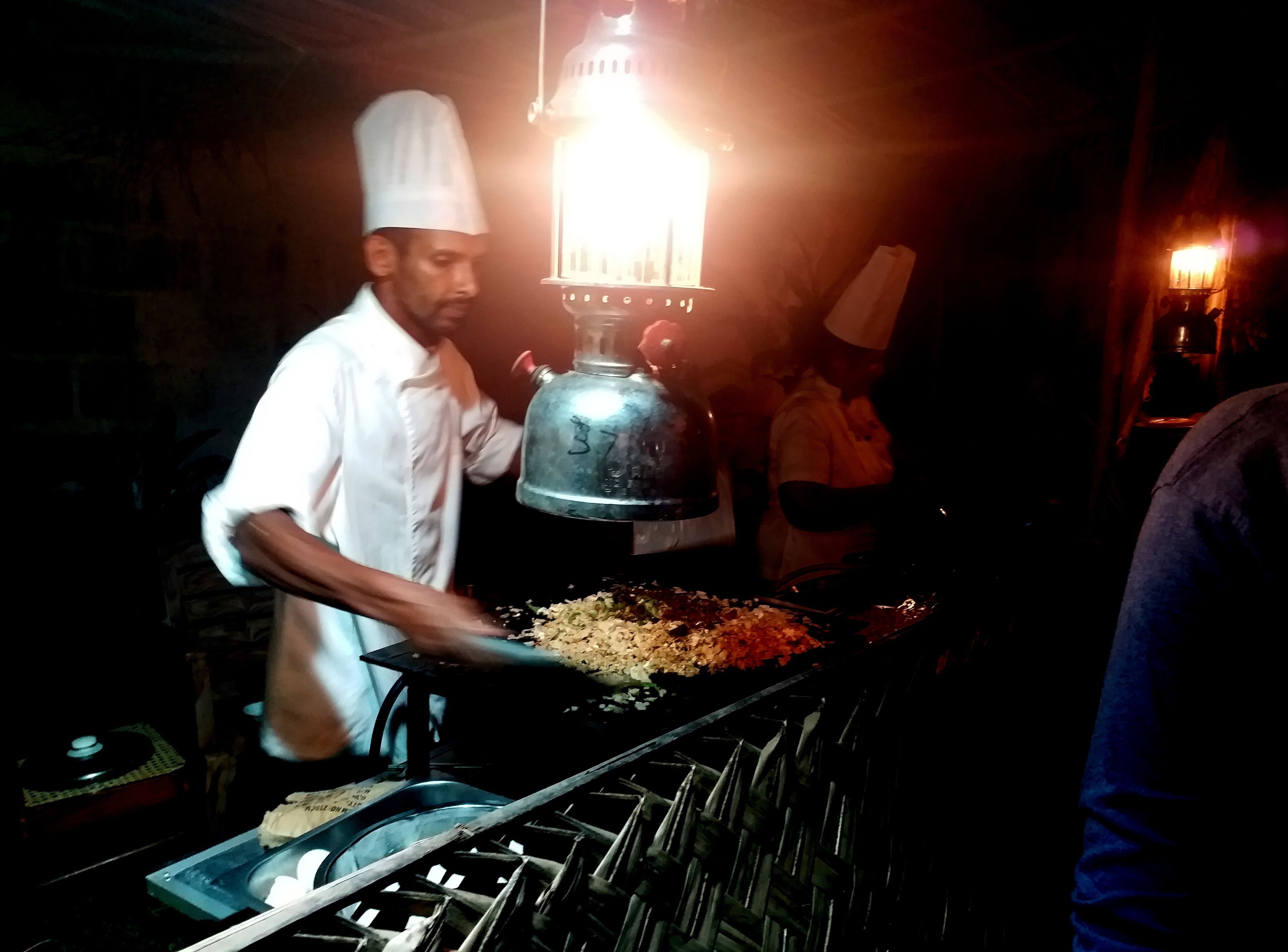 A chef preparing food in a dimly lit kitchen with a large stove and a hanging light fixture.