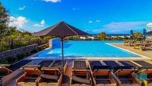 A poolside area with lounge chairs and umbrellas, a clear blue sky, and a view of the ocean.
