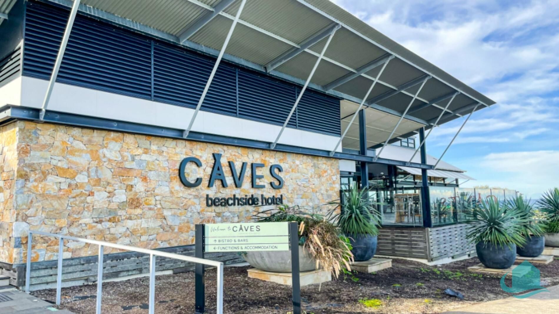 L’extérieur de l’hôtel Caves Beachside se distingue par sa façade en pierre, son entrée couverte et une enseigne indiquant l’emplacement du bar et des salles de réception.
