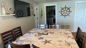 Un coin repas avec une table recouverte d’une nappe à motifs marins, des chaises en bois, un téléviseur fixé au mur et des éléments de décoration murale, dont une roue de navire.