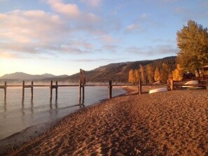 A beach with a wooden pier extending into calm waters, surrounded by trees and mountains in the background.