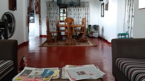 A living room with a wooden dining table, chairs, a television, and newspapers on the floor. - Nature Villa 107 Kandy - Hostel (Kandy)