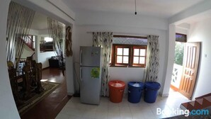 A kitchen with a refrigerator, two trash bins, a window with curtains, and a door leading to another room. - Nature Villa 107 Kandy - Hostel (Kandy)